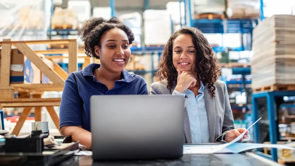 Two employees review online training and learning course on laptop on shop floor.