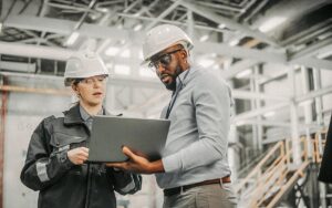 Two Diverse Professional Heavy Industry Engineers Wearing Safety Uniform And Hard Hats Working On Laptop Computer. African American Technician And Female Worker Talking On A Meeting In A Factory.