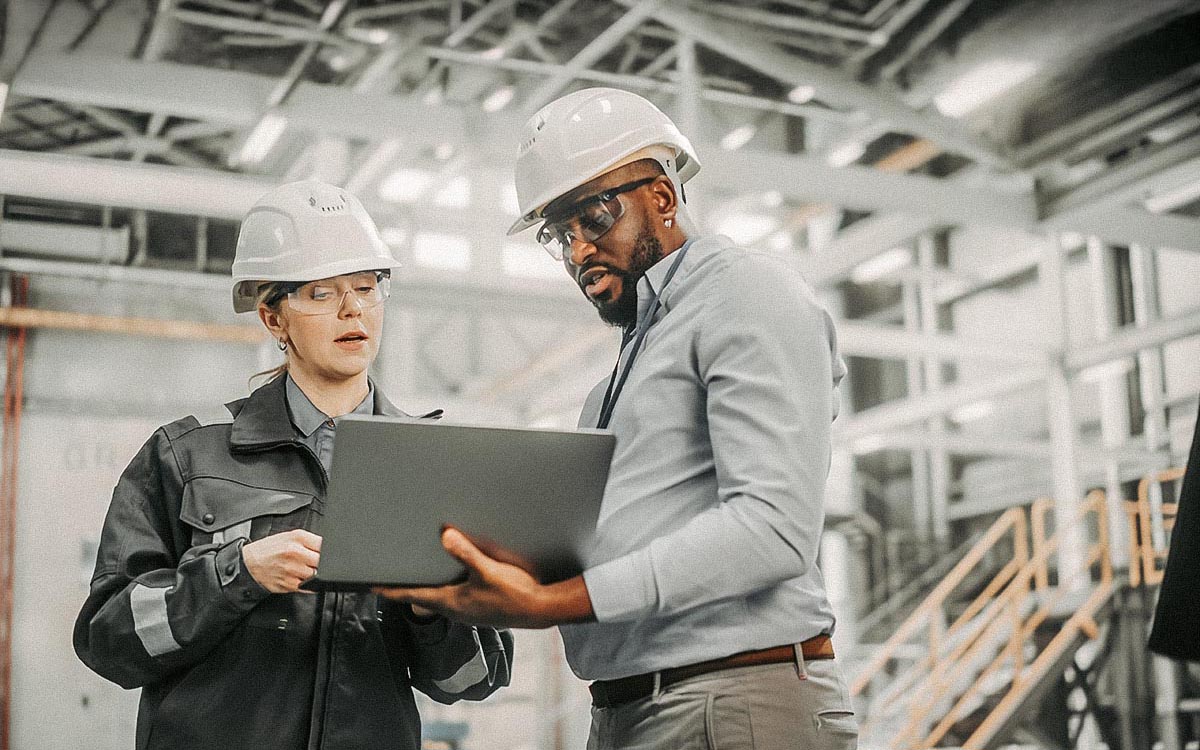 Two Diverse Professional Heavy Industry Engineers Wearing Safety Uniform And Hard Hats Working On Laptop Computer. African American Technician And Female Worker Talking On A Meeting In A Factory.