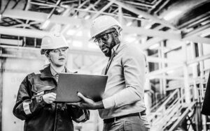 Two Diverse Professional Heavy Industry Engineers Wearing Safety Uniform And Hard Hats Working On Laptop Computer. African American Technician And Female Worker Talking On A Meeting In A Factory.