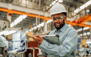 Male African Maintenance Engineer Working In Automotive Parts Production Line While Using A Digital Tablet For Check About A Data Reviews .he 'd Tried To Manage And Balance Process Line.