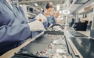 Close Up Of A Female Electronics Factory Worker In Blue Work Coat Assembling Laptop's Motherboard With A Screwdriver. High Tech Factory Facility With Multiple Employees.