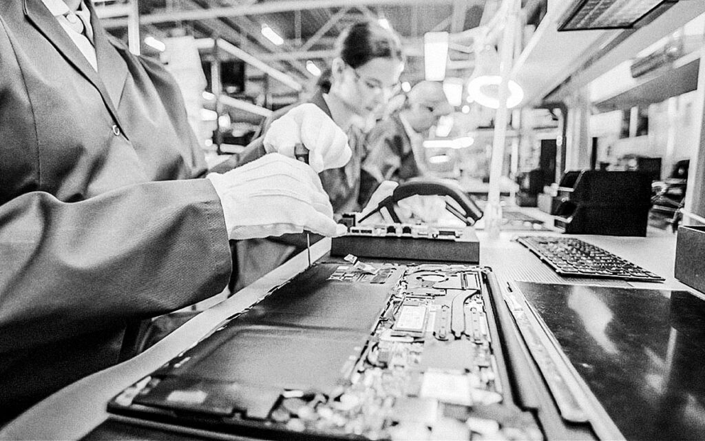 Close Up Of A Female Electronics Factory Worker In Blue Work Coat Assembling Laptop's Motherboard With A Screwdriver. High Tech Factory Facility With Multiple Employees.