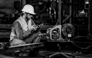Worker Handles Metal At Lathe Turner Measures The Dimensions Of The Metal Workpiece With A Caliper In Uniform With Safety. Work On A Lathe.