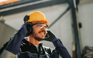 A Cheerful Male Worker Wearing A Safety Helmet And Protective Gear Speaks On A Headset In A Bustling Factory Environment, Emphasizing Industry Safety And Productivity.