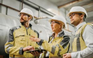 Three Heavy Industry Engineers Stand In Pipe Manufacturing Factory, Use Digital Tablet Computer, Have Discussion. Large Pipe Assembled. Design And Construction Of Oil, Gas And Fuels Transport Pipeline