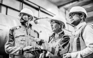 Three Heavy Industry Engineers Stand In Pipe Manufacturing Factory, Use Digital Tablet Computer, Have Discussion. Large Pipe Assembled. Design And Construction Of Oil, Gas And Fuels Transport Pipeline