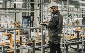 Car Factory Engineer In Work Uniform Using Laptop Computer. Automotive Industrial Manufacturing Facility Working On Vehicle Production With Robotic Arms Technology. Automated Assembly Plant.