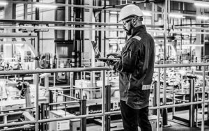 Car Factory Engineer In Work Uniform Using Laptop Computer. Automotive Industrial Manufacturing Facility Working On Vehicle Production With Robotic Arms Technology. Automated Assembly Plant.